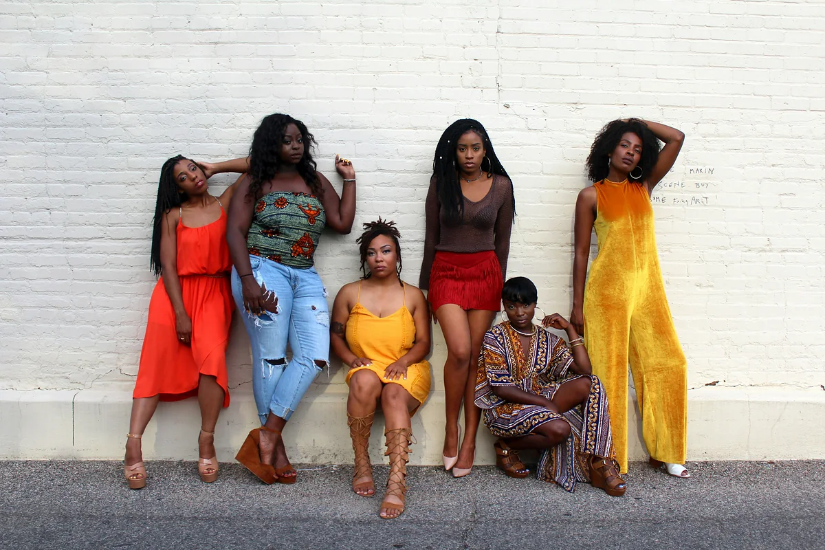 Six women in colourful clothes gathered against a white brick wall