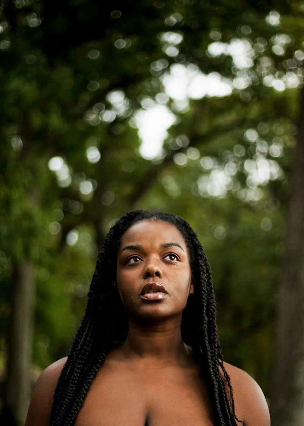 A woman with long braids looking upward, surrounded by trees